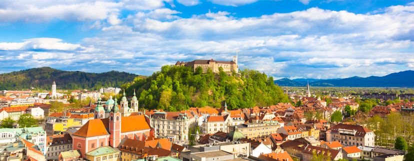 De stad Ljubljana met in het midden het kasteel op een heuvel en op de achtergrond bergen en een blauwe lucht met wolken