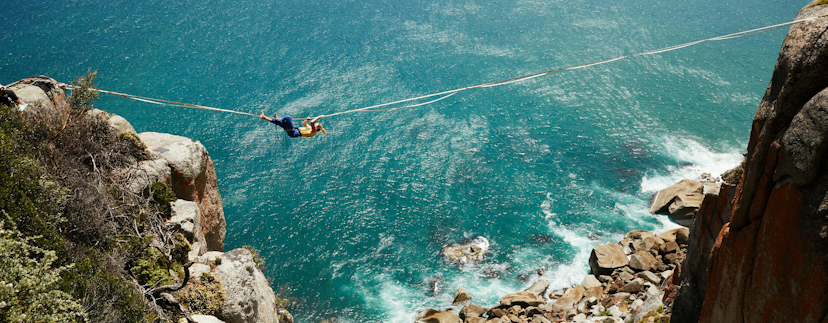 Een vrouw die tussen twee kliffen naast de zee aan een touw hangt. Onder haar liggen losse kleine rotsen in de zee.