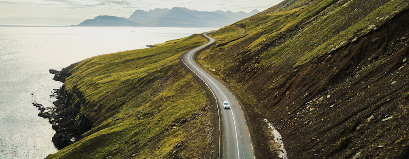 Een grijze auto die over een slingerende weg rijdt in IJsland. De weg loopt over een heuvel die in de zee uitkomt. Het landschap is een mix van gras en aarde. Op de achtergrond zijn mistige bergen te zien.