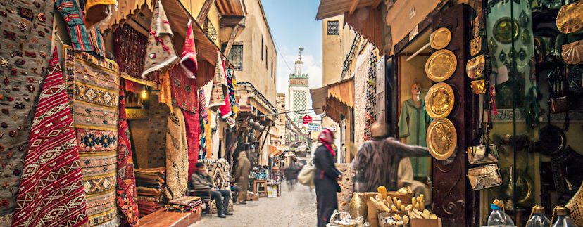 De medina van Fez, met een aantal mensen op de straat. Links hangen veel gekleurde kleden, en rechts meerdere gouden schalen, flessen en andere waren in winkels.