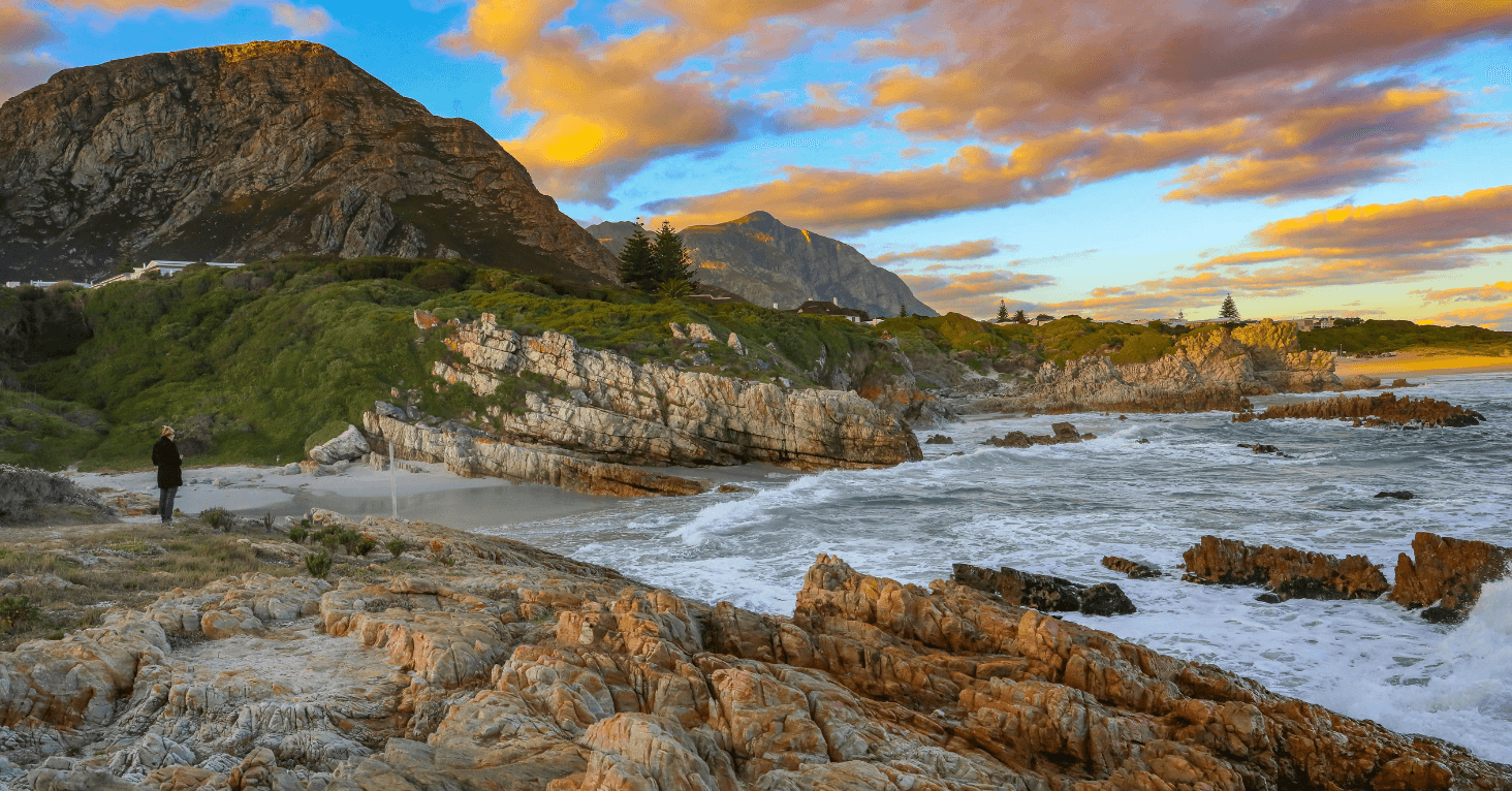 Coastal landscape with rocky shoreline, green hills, and a mountain backdrop under a vibrant sunset sky. Waves crash on the rocks.