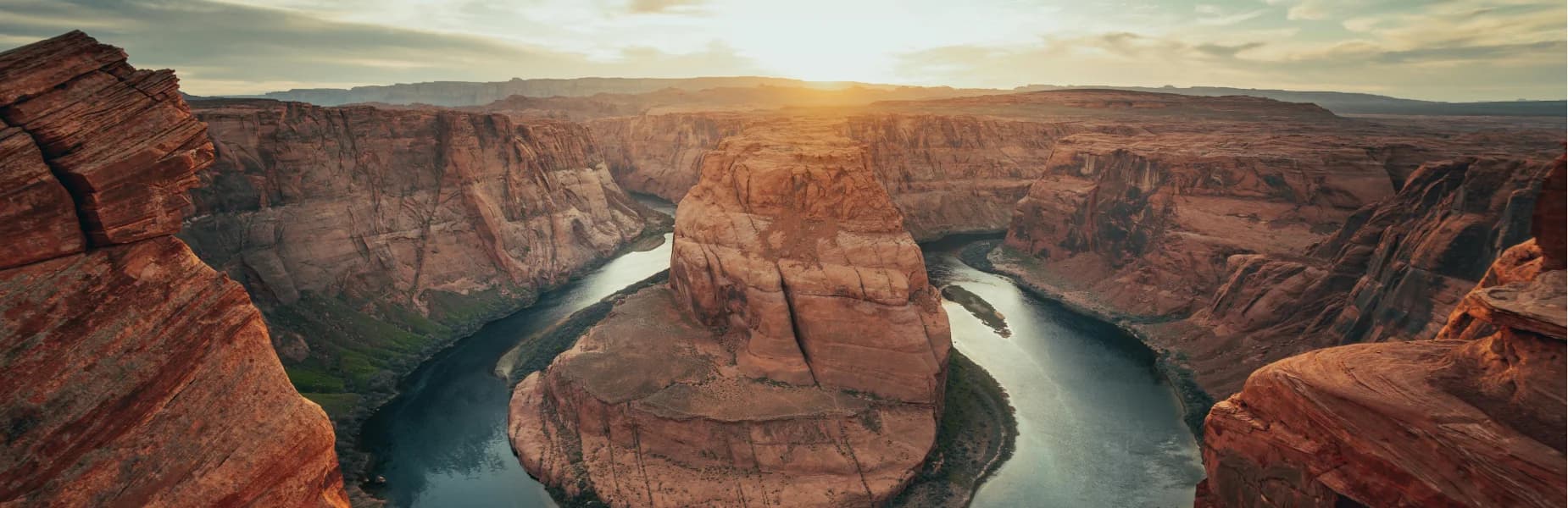 Majestic canyon landscape at sunset, with a river curving around a large rock formation, surrounded by steep, rugged cliffs.