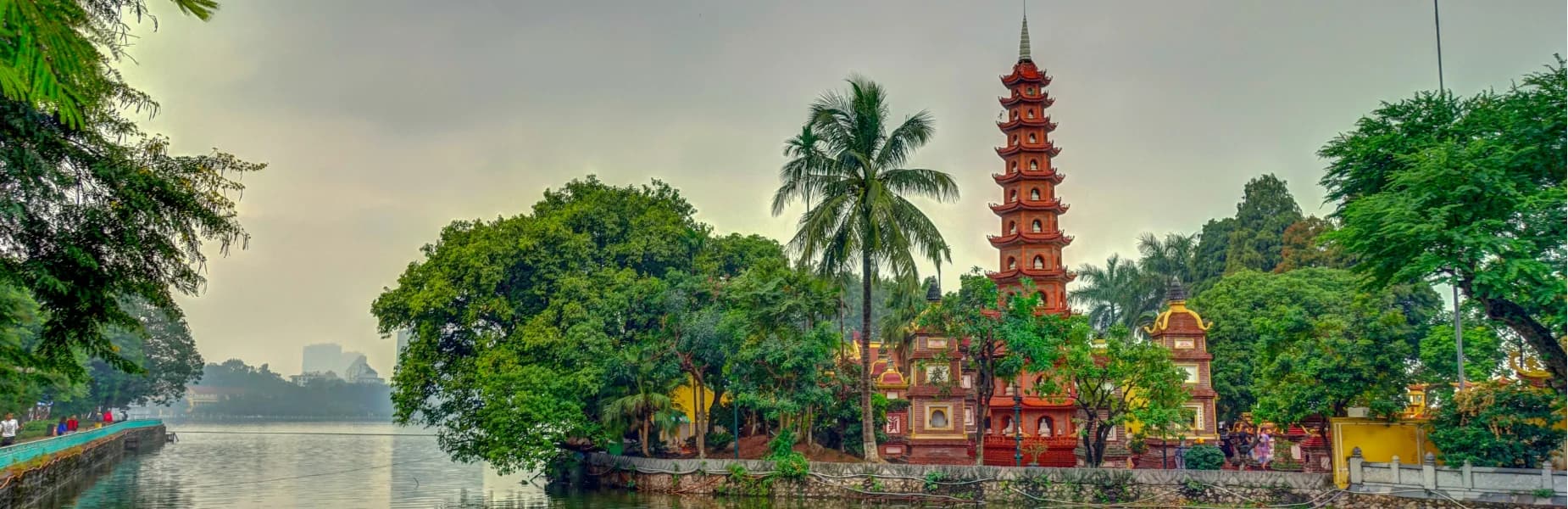 A tall pagoda set amidst lush greenery by a serene lake, with city buildings faintly visible in the misty background.