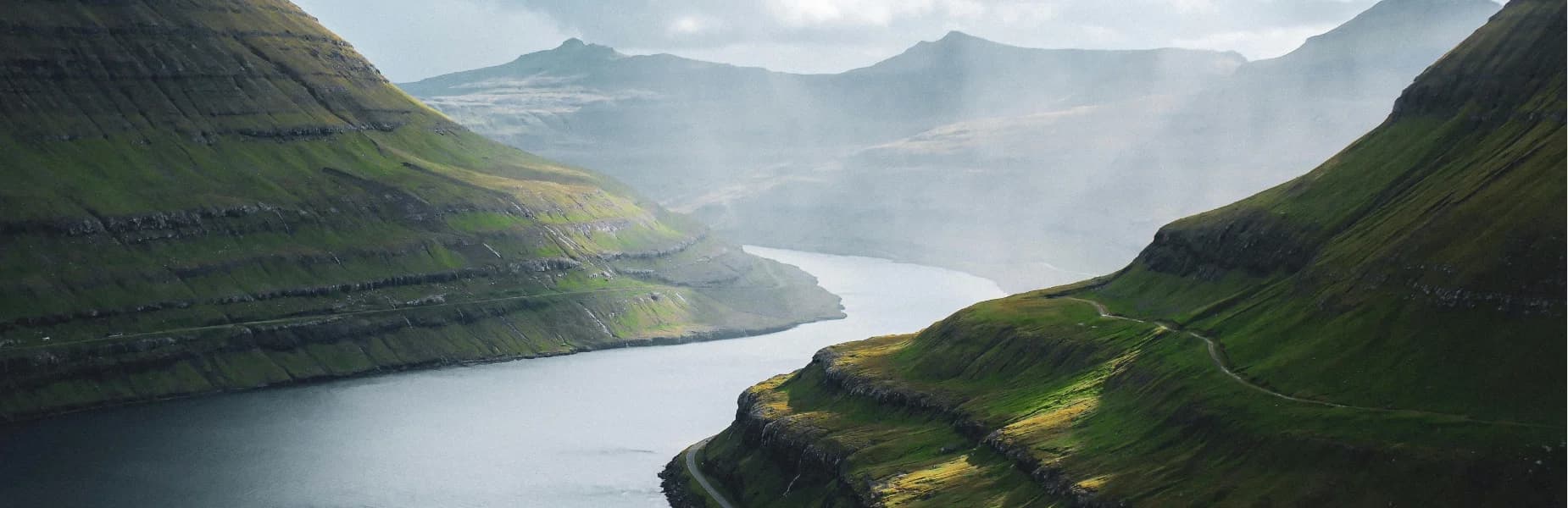 A serene landscape of green, grassy hills and a winding, calm river under a misty sky with distant mountain silhouettes.