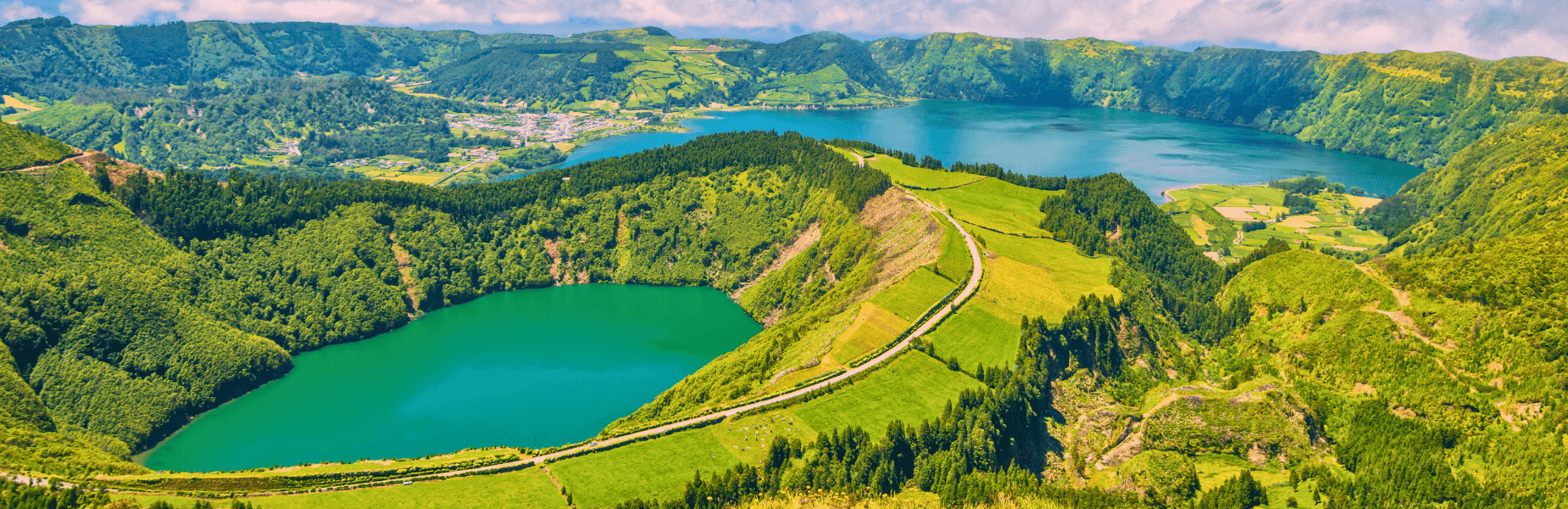 Aerial view of lush, green hills surrounding two scenic crater lakes with a town and winding roads, under a partly cloudy sky.