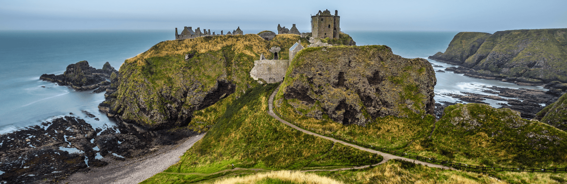 Scenic view of Dunnottar Castle perched on a rocky cliff overlooking the sea, surrounded by green grass and rugged coastline under a cloudy sky.