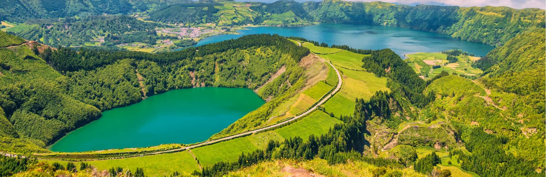A panoramic view of two vibrant blue lakes surrounded by lush green mountains and fields, under a clear blue sky.