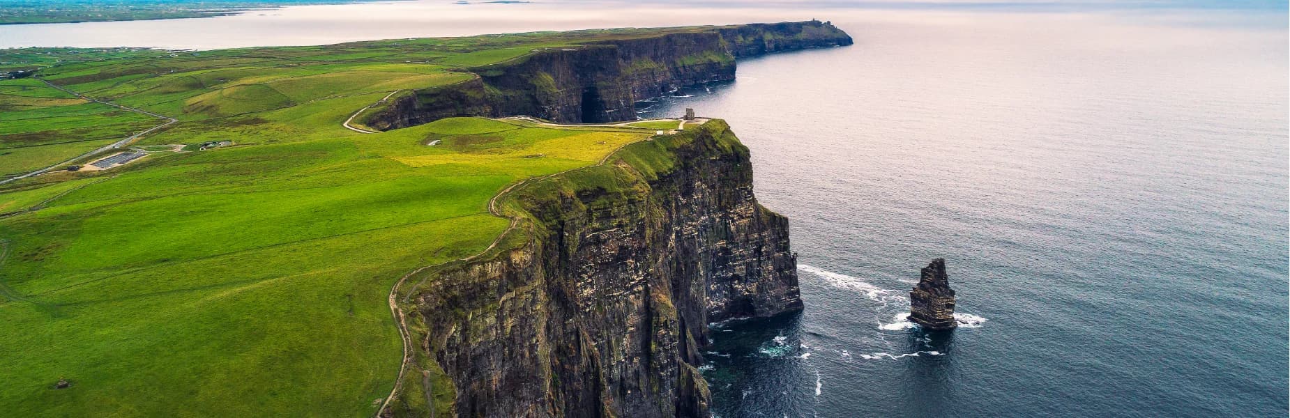 Aerial view of dramatic coastal cliffs with lush green fields atop, adjacent to a calm ocean, under a light, cloudy sky.