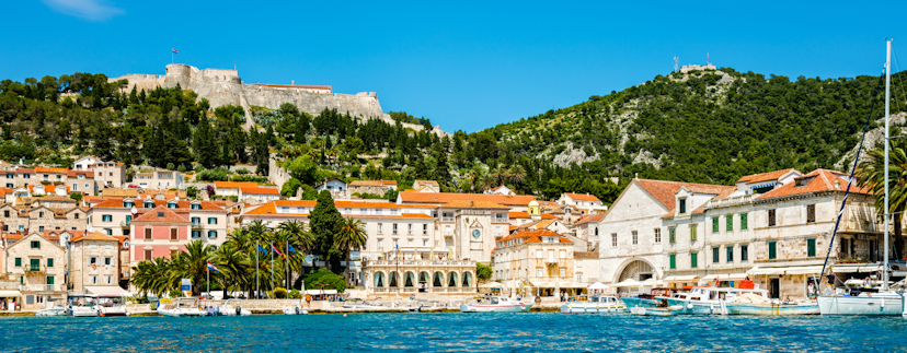 Op de voorgrond de diepblauwe zee met kleine golven. In de mediterraanse haven liggen enkele luxe boten. Aan de kust typisch Kroatische huizen met licht steen en rode daken, en palmbomen aan de boulevard. Boven de huizen uit een berg met bomen, en bovenin onder de blauwe lucht een fort op de berg.