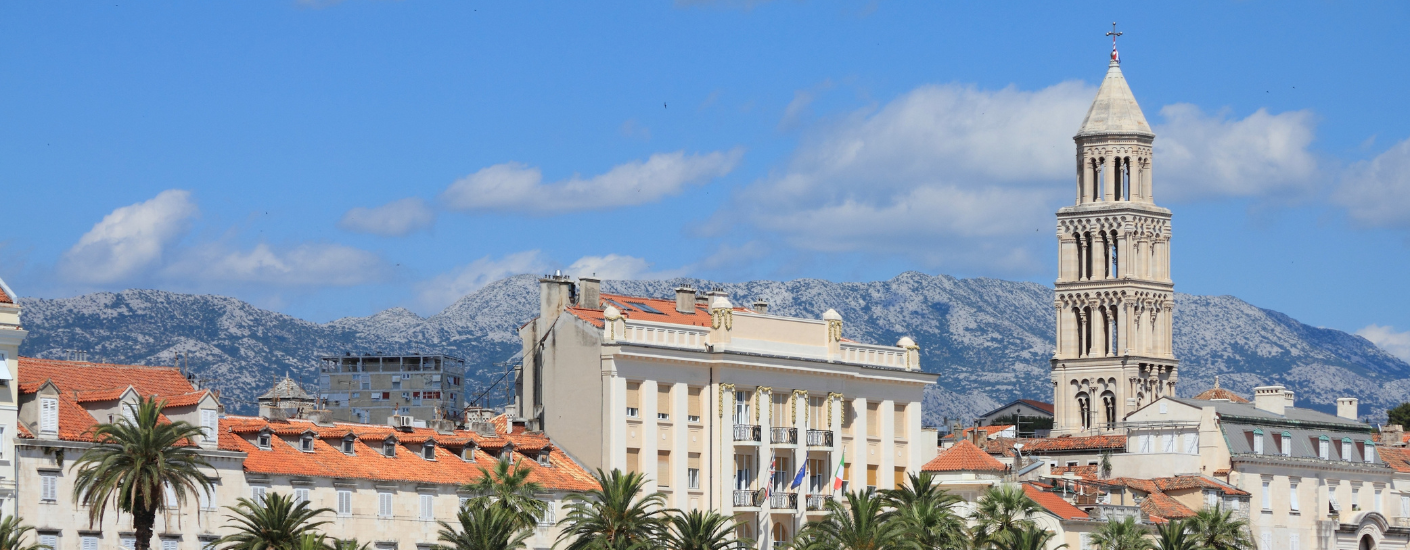 De toren van het paleis in Split, en de huizen in split met de bergen op de achtergrond. Blauwe lucht met een paar wolken, en palmbomen op de voorgrond.