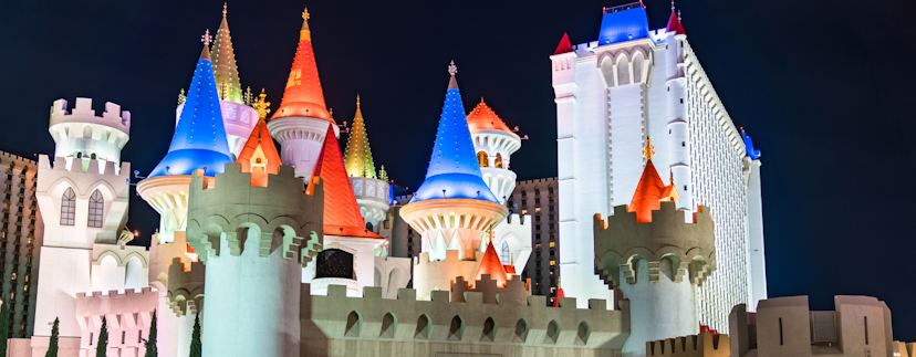 Het Excalibur Hotel in Las Vegas. Verschillende torens en kasteelmuren met kantelen vormen een Disney-achtig kasteel hotel. Daken zijn wit en steenkleurig of blauw, rood, geel en paars verlicht. Op de achtergrond is een groter, flatgebouw-achtig deel van het hotel te zien. De lucht is donker.
