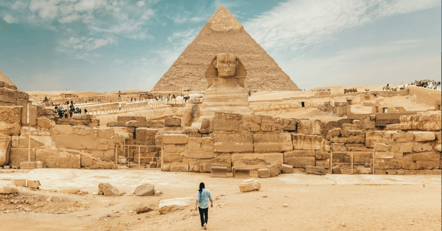 A person walks towards the Sphinx and Great Pyramid of Giza under a clear blue sky, with tourists gathered in the background.