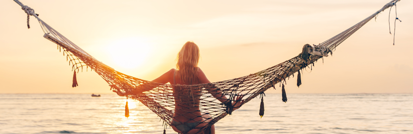 Een blonde vrouw van de achterkant in een hangmat kijkend naar de zee met zonsondergang