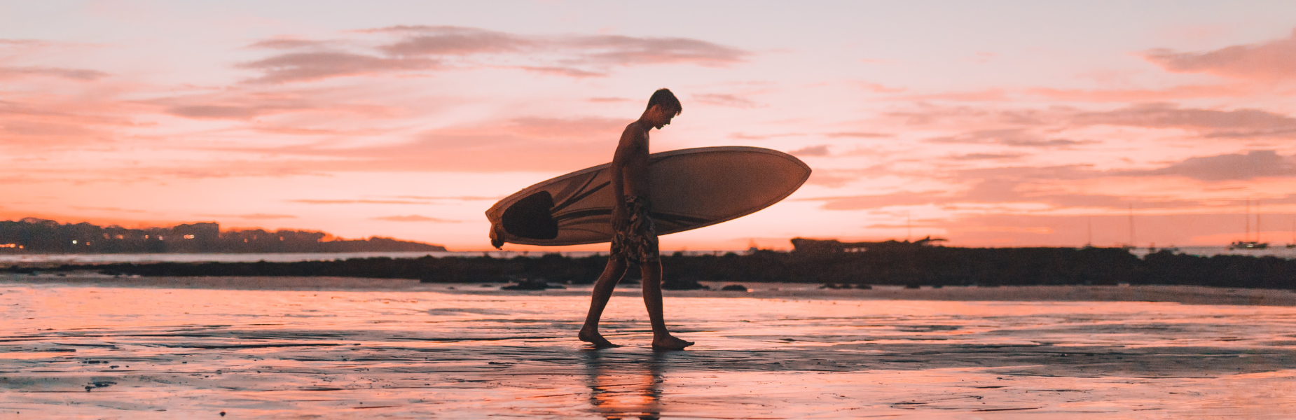 Een surfer met een surfboard die langs de zee loopt. Op de achtergrond zie je water, de ondergaande zon en een rode lucht met wolken.