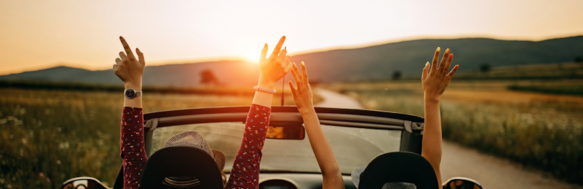 Meisjes met armen in de lucht in een auto met open dak en zonsondergang achter de bergen